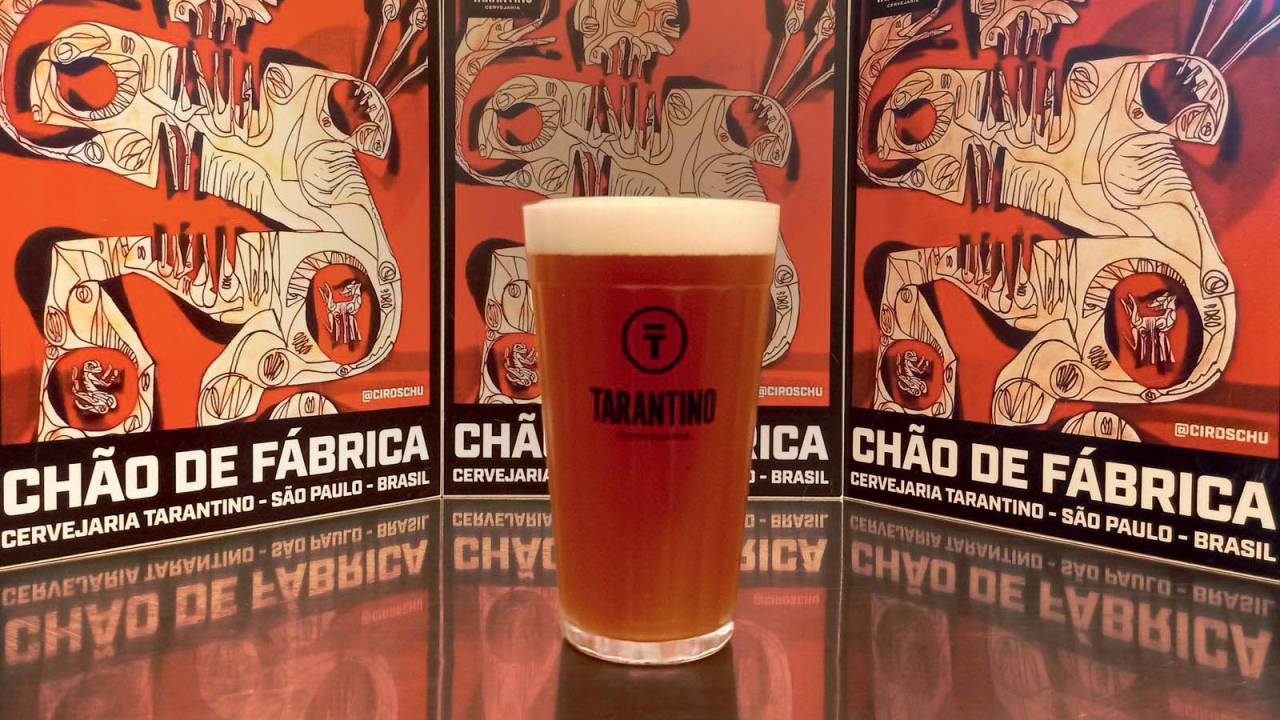 Copo de cerveja em que se lê Tarantino sobre mesa espelhada com três banners ao fundo onde se lê Chão de Fábrica