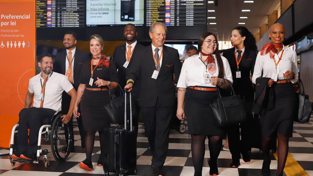 Equipe de voo anda por corredor de aeroporto vestindo uniforme e tênis.