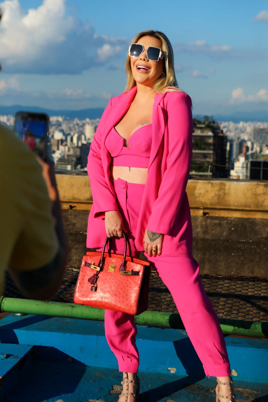 Kamilla Agacci Boing (Divulgação) Mulher branca e loira posa no topo de prédio com roupas rosa pink e bolsa na mão direita.
