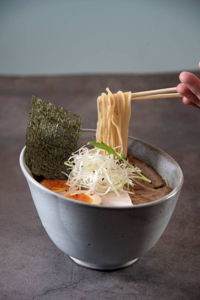 Tigela com lamen alga nori ovo e copa lombo, com mão pegando com hashi um pouco de macarrão