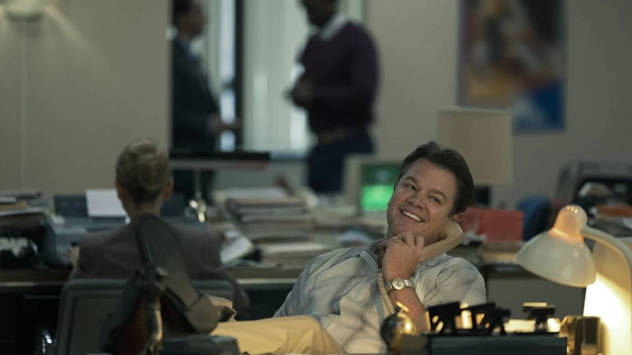 Matt Damon, com pés para cima da mesa, sorri no telefone