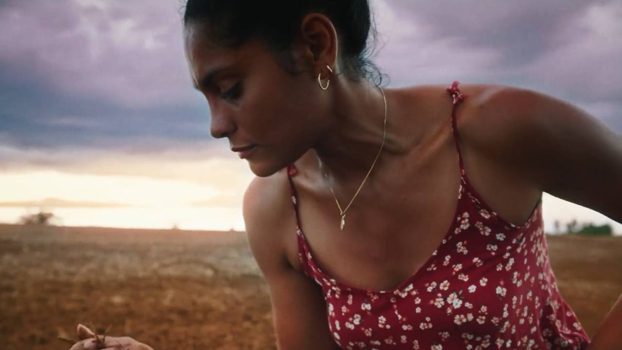 Mulher negra de cabelos presos em coque veste roupa vermelha com flores brancas de alças e está ajoelhada em trecho de fazenda com chão de terra