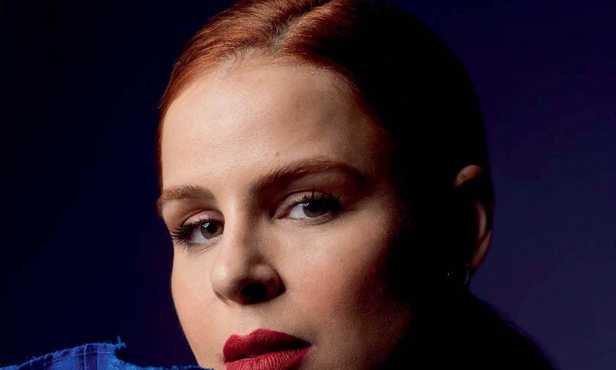 Mulher branca de coque e batom vermelho encara a câmera em local de fundo azul e roupa com babados azuis próximos ao rosto.