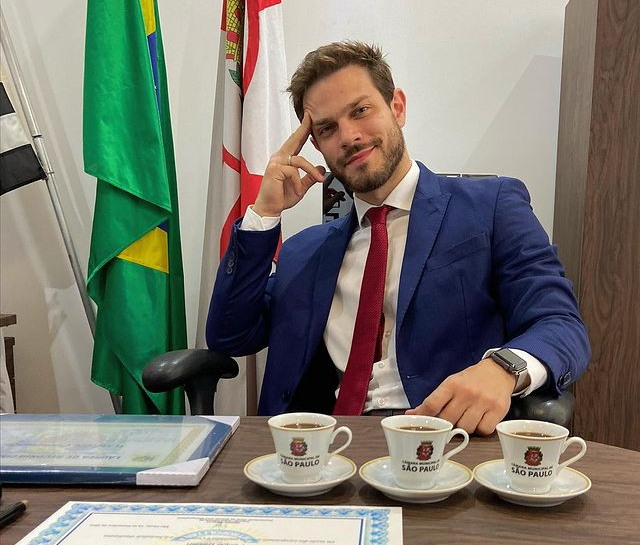 homem loiro de terno azul sentando à mesa com três xícaras de café em sua frente