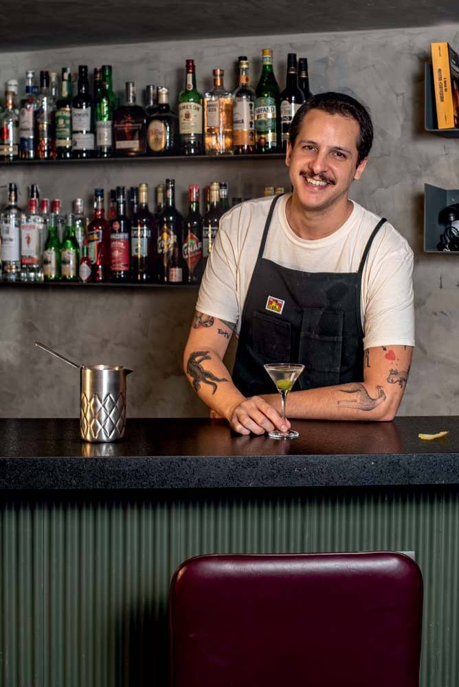 Bargo Mooca Homem branco sorrindo atrás de balcão de bar segurando um dry martini.