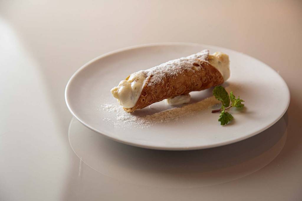 Cannolo recheado de creme de ricota com açúcar de confeiteiro por cima em prato de louça branca.