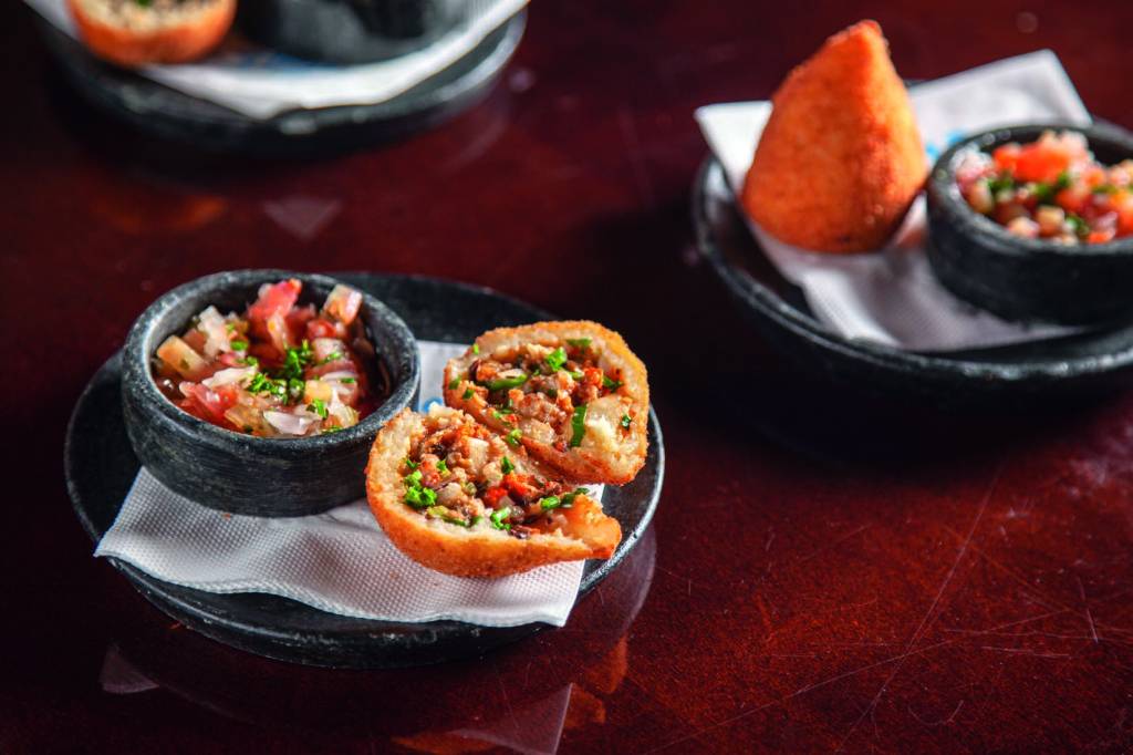Dia das Mães: Rancho Jeri – coxinhas de frutos do mar Coxinha cortada ao meio recheada de frutos do mar sobre prato de pedra ao lado de potinho de vinagrete em mesa de madeira.