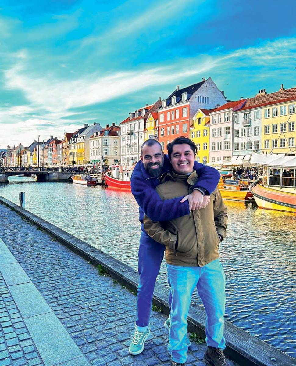 Enrico Andrade e Rafael Brito – Copenhagen, Dinamarca (Arquivo Pessoal).jpeg Casal de homens brancos se abraça em frente a rio com casinhas coloridas ao fundo. Vestem roupas de frio com calça jeans.