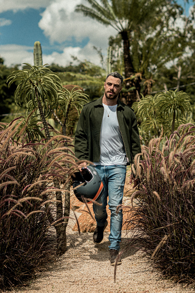 Felipe Titto (Viviane Dente-Divulgação) (3).jpg Homem branco de barba anda no meio de uma plantação com jardim ao fundo. Veste jaqueta verde, camiseta clara e jeans clara rasgada. Está segurando um capacete de moto.
