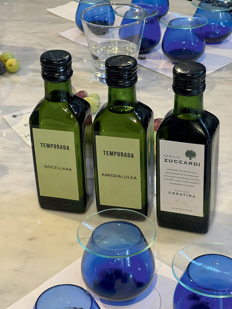 Azeite – Família Zuccardi Três garrafas verdes de azeite lado a lado junto e taças azuis.