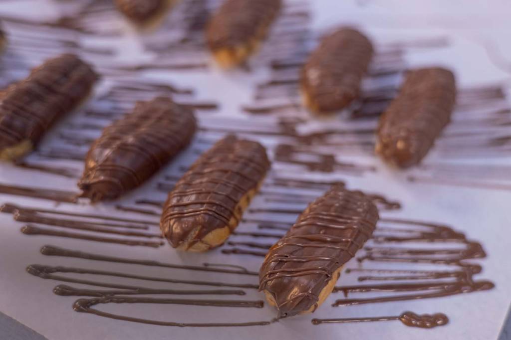 Mini eclairs alinhadas em diagonal sobre tampo branco "manchado" de linhas de chocolate.