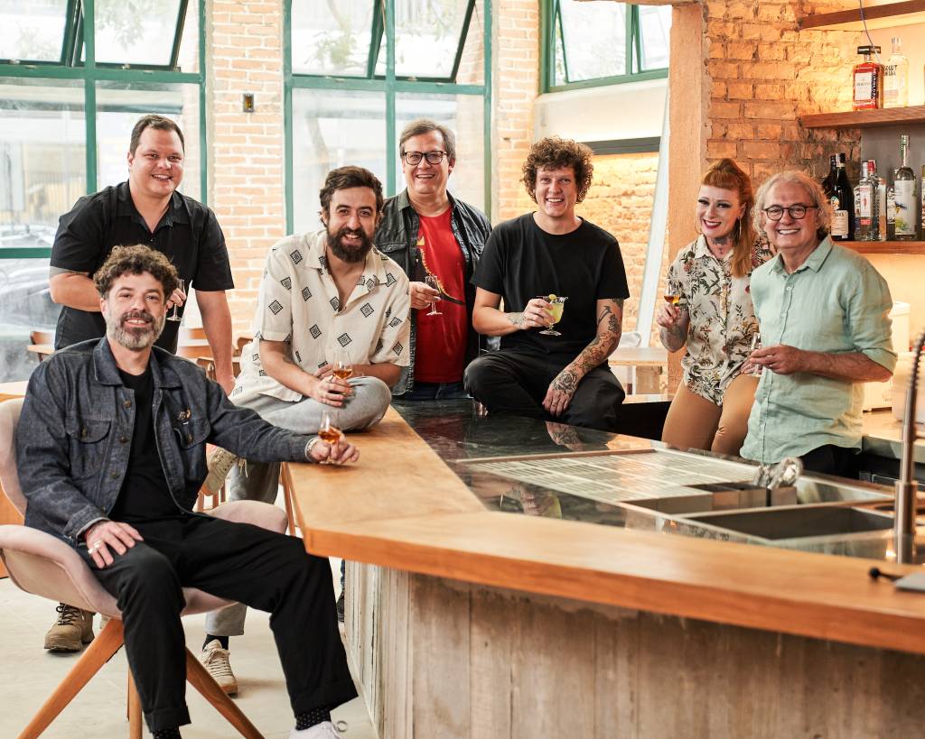 Seis homens brancos e uma mulher branca sentados no balcão de um bar olhando para a foto.