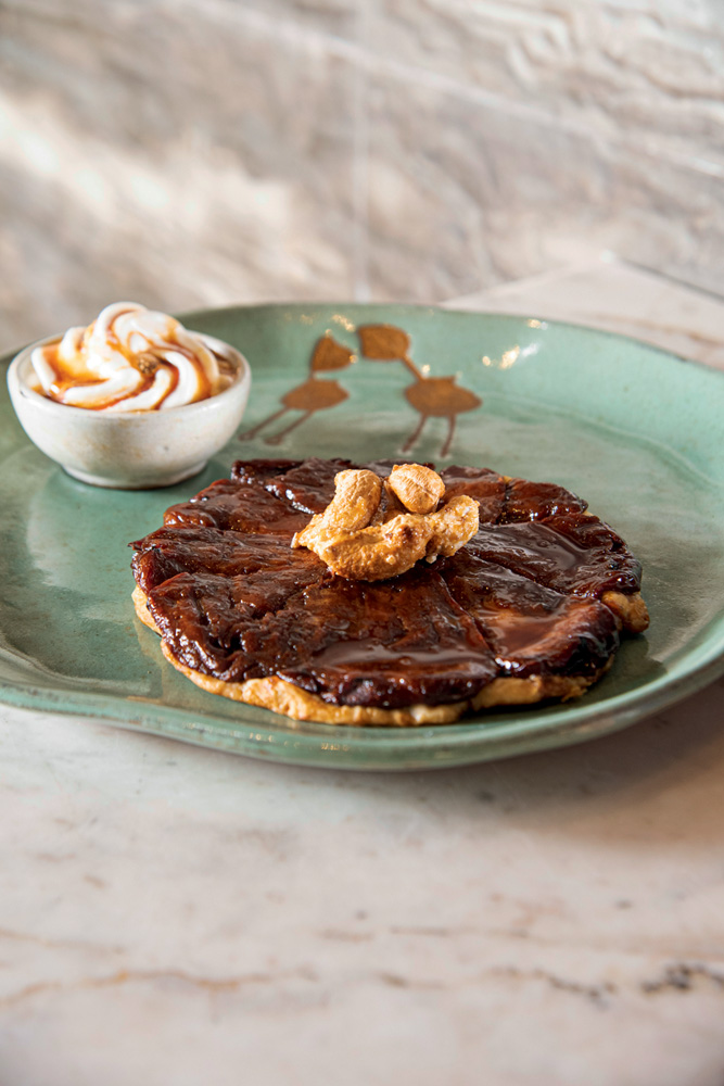 Tarte tatin – Taraz Prato de cerâmica verde com torta doce achatada coberta por castanha-de-caju caramelada ao lado de potinho com sorvete.