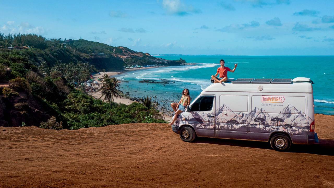 Foto de casal em cima de motorhome e praia de mar verde ao fundo.