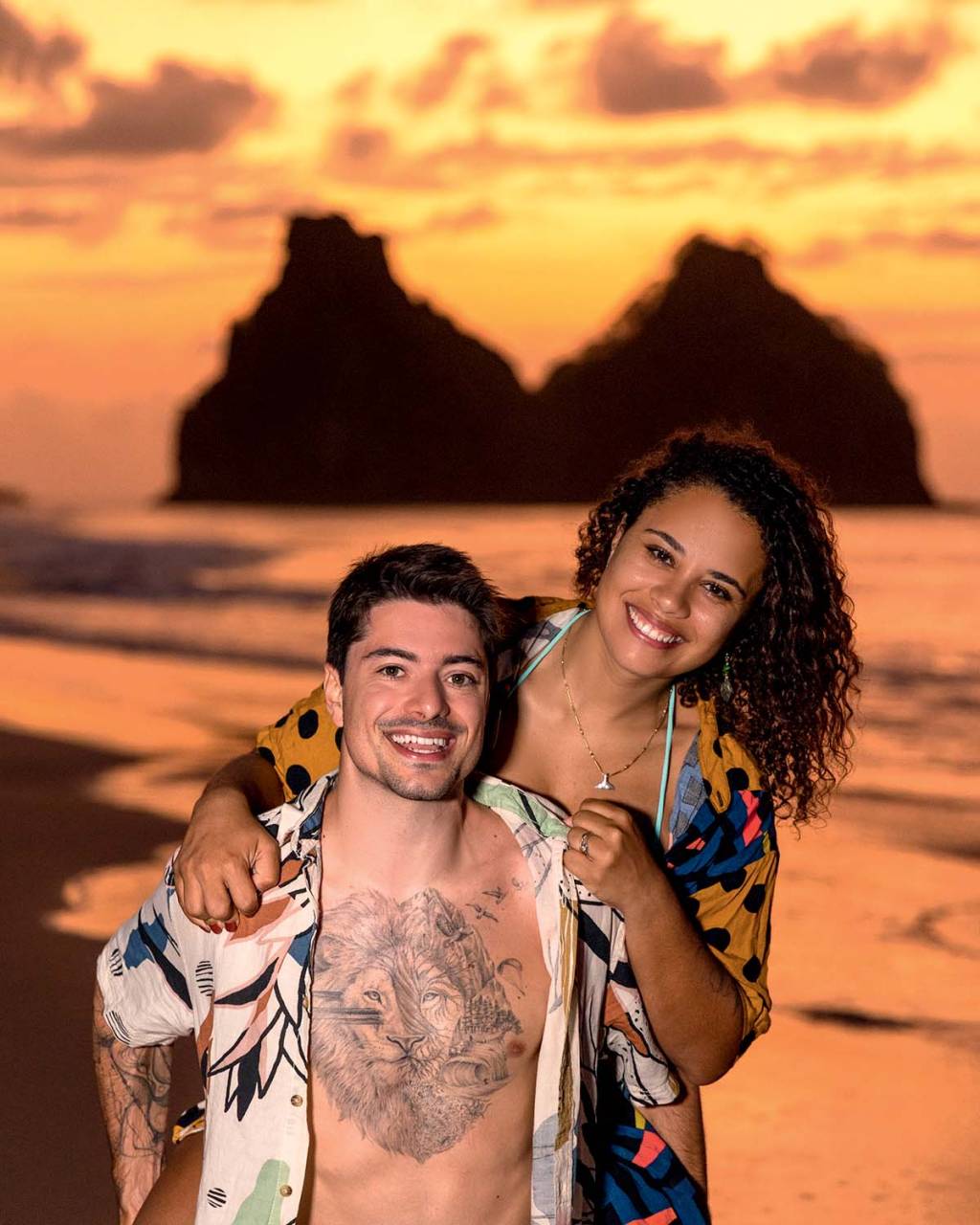 Traveleiros (Divulgação) (2).jpg Homem branco de peito tatuado e camisa estampada posa com a esposa segurando seus ombros, uma mulher negra de cabelos cacheados. Posam com uma praia em tons alaranjados ao fundo.