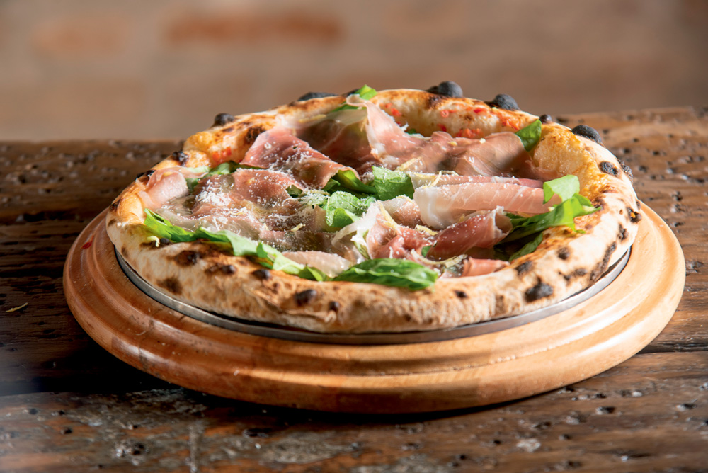 Villa Napoli Pizza individual de bordas altas com presunto cru, parmesão, rúcula e raspas de limão-siciliano sobre madeira.