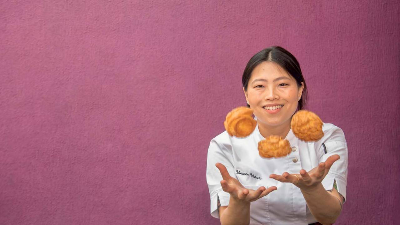 Mulher asiática de dólmã em frente à parede roxa jogando três doces choux para o alto.