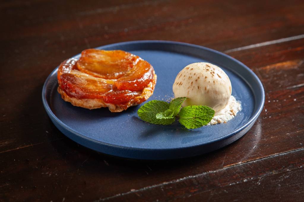 Tarte tatin – Avec Gastronomie Prato azul sobre mesa de madeira com tarte tatin mini ao lado de bola de sorvete de creme enfeitada com ramo de hortelã.