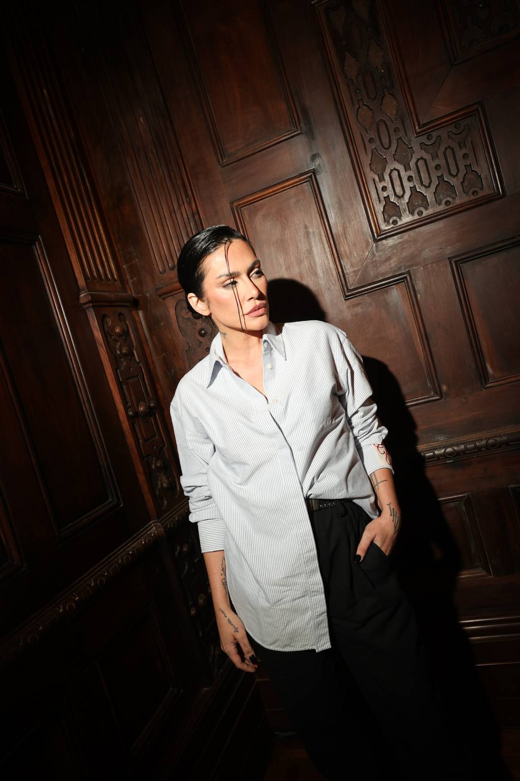 Cléo Pires Mulher branca de cabelo preto preso para trás, com uma tira em frente ao rosto, posa com uma mão no bolso da calça preta e veste camisa larga clara.