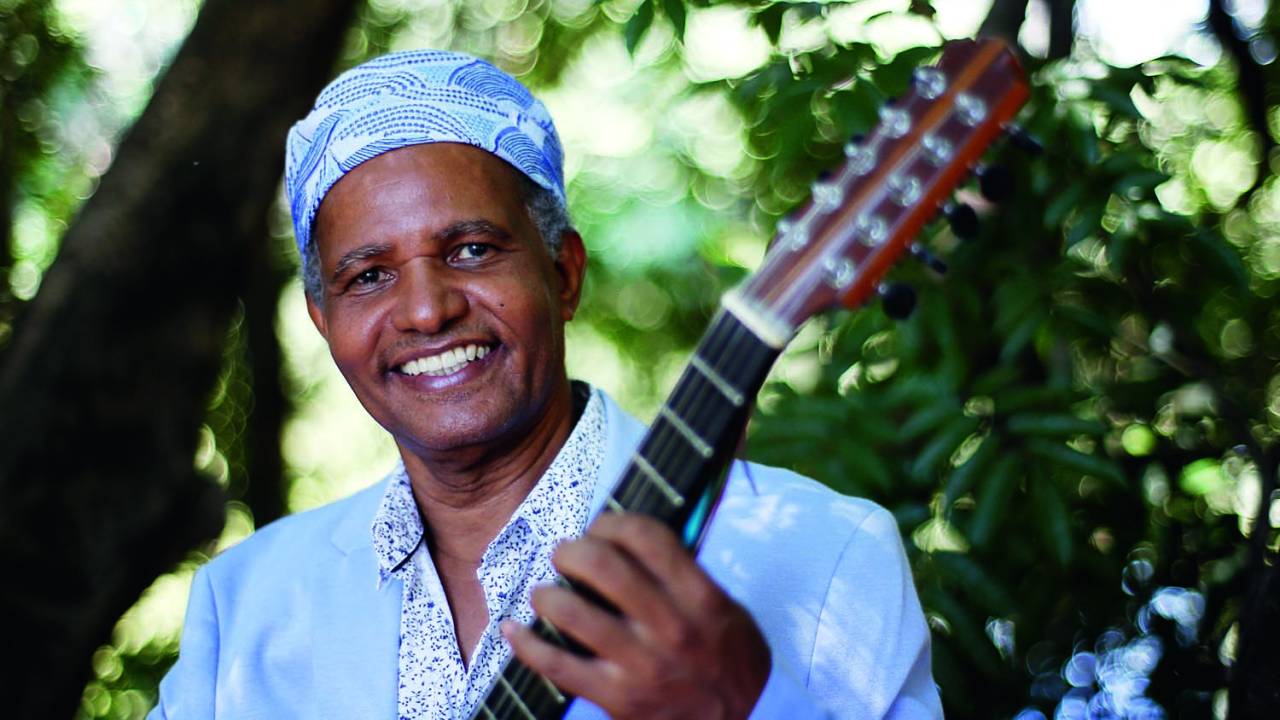Imagem mostra homem com roupa azul, segurando viola, sorrindo