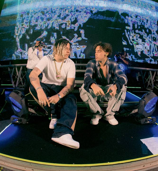 Imagem mostra dois homens sentados em palco, se olhando