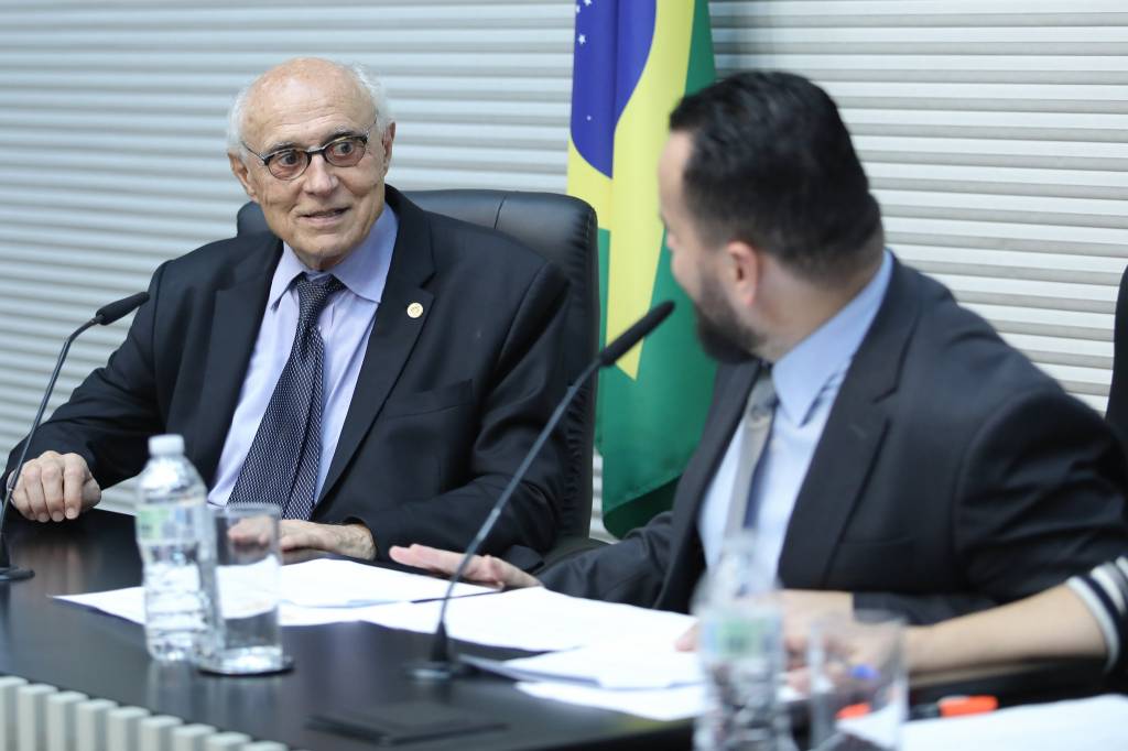 Um homem grisalho conversa com um homem de cabelo castanho em uma mesa. Atrás, a bandeira do Brasil.