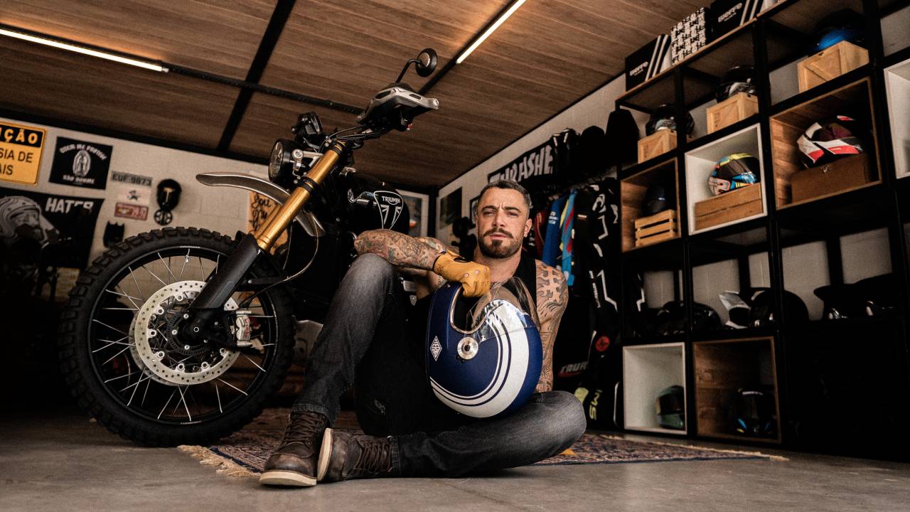 Homem branco de barba posa sentado no chão com capacete no colo e moto e prateleiras ao fundo.