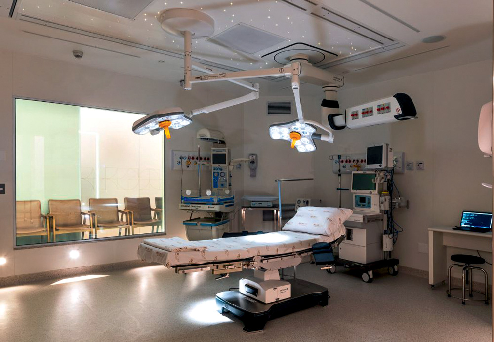 Imagem de quarto hospitalar com grande janela, maca e equipamentos no teto.