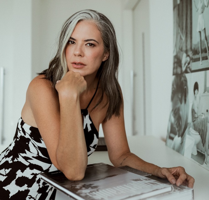 Mulher de cabelos lisos com franja grisalha encara a câmera com uma mão no queixo. Veste vestido preto e branco e está apoiada em mesa com um livor aberto.