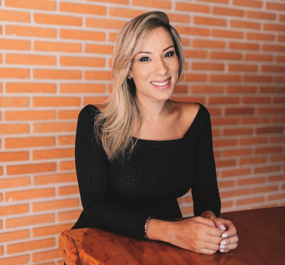 Mulher loira posa sorrindo sentada atrás de mesa de madeira, os antebraços apoiados e unidos. Veste camiseta de manga longa preta. Ao fundo, parede de ladrilhos.