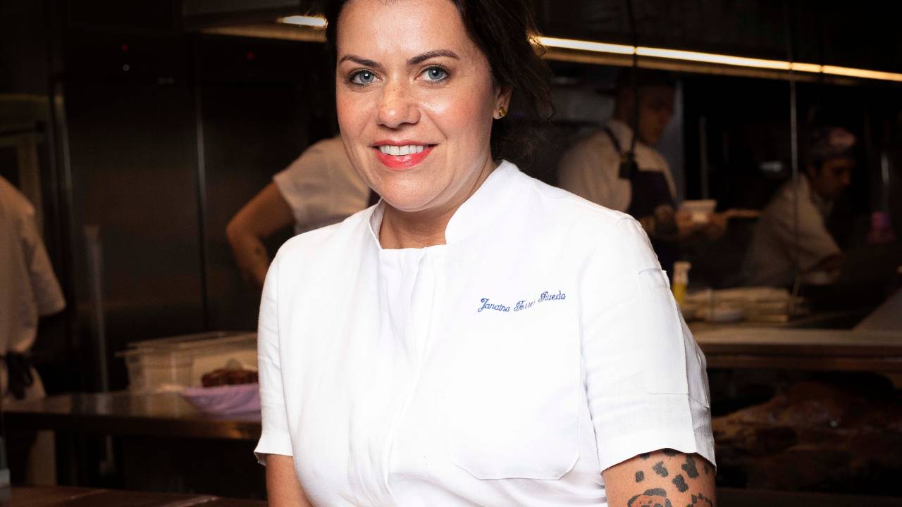 Mulher de olhos claros e cabelos escuros posa com um dos braços tatuados em destaque, apoiada em balcão de cozinha profissional. Veste dolmã branca.
