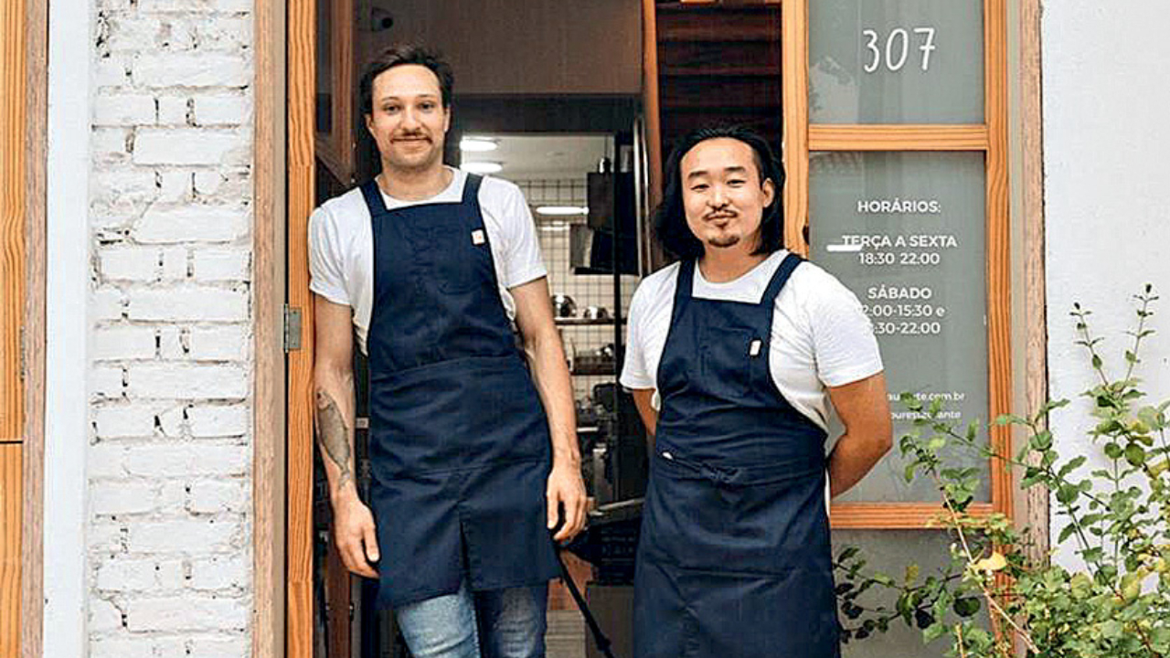 Dois homens, um branco à esquerda e outro asiático à direita, postados usando aventais em frente a restaurante.