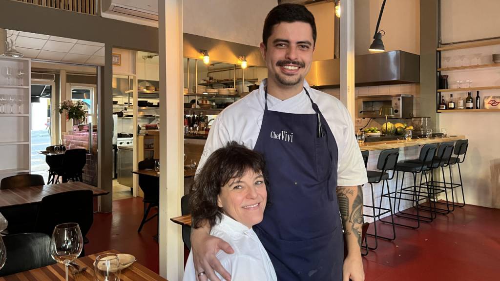 Chef Viviane Gonçalves de dólmã sentada abração subchef homem em pé de avental.