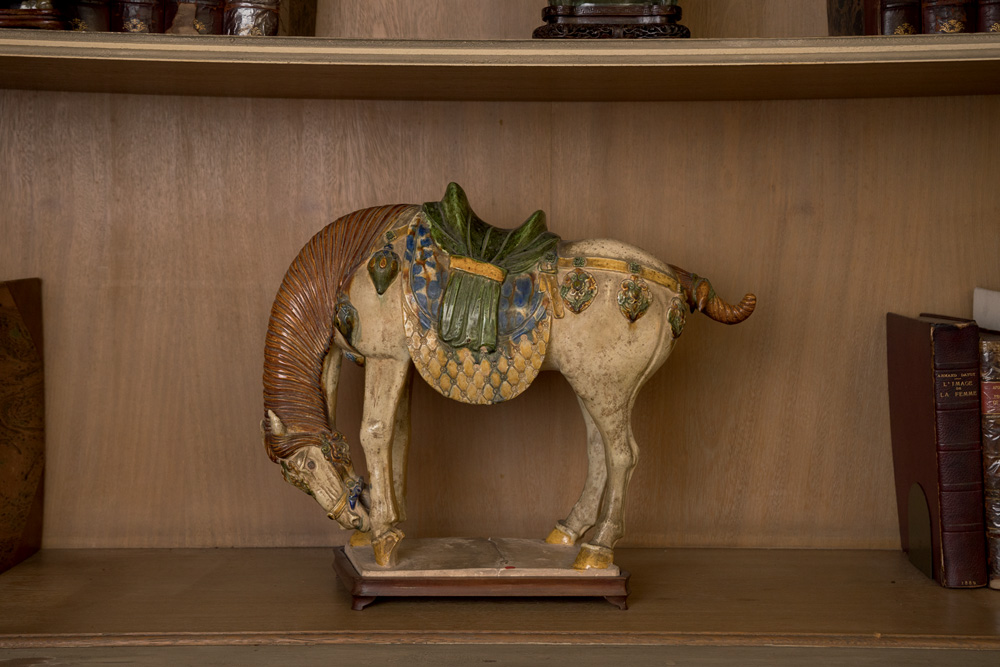 PÁGINA 5 – Leilão Familia Prado – Cavalo cerâmica dinastia Tang (600-900 DC) (Wanezza Soares-Veja SP) (8) Cavalo de porcelana aparece de lado, com a cabeça abaixada, em prateleira de madeira.