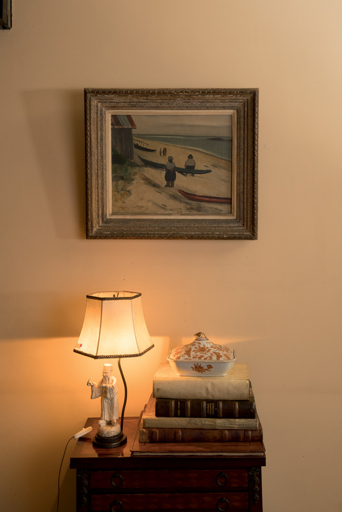 Leilão Familia Prado (Wanezza Soares-Veja SP) (7) Parede com quadro de paisagem e uma mesa de canto com livros e abajur de marfim.