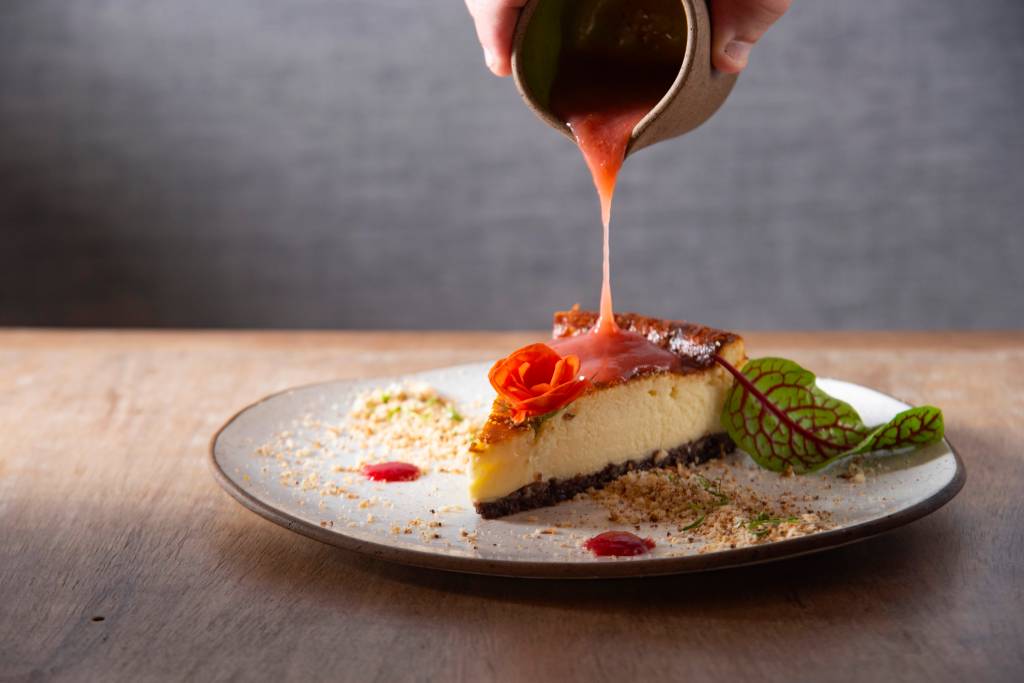 Paseo Cocina y Tal Prato de cerâmica com fatia de torta de queijo sendo derrubada geleia de figo de um potinho por cima.