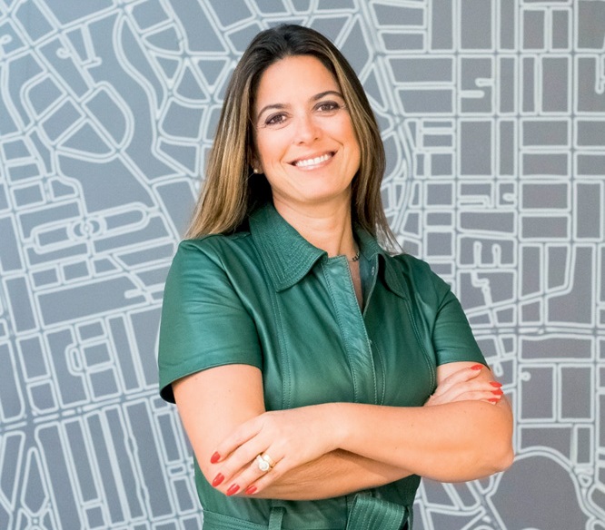 Mulher branca de cabelos lisos castanhos posa de braços cruzados em fundo com linhas brancas e fundo cinza claro. Veste vestido verde com gola e mangas curtas.