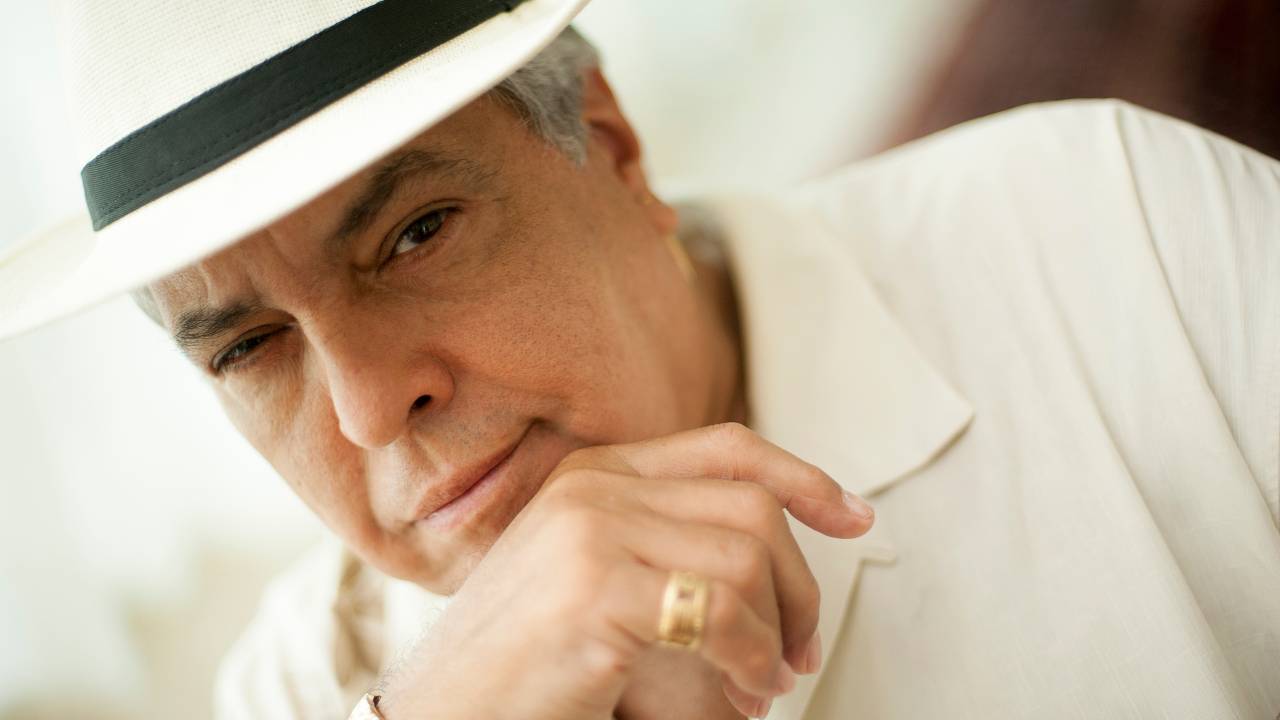 Homem branco grisalho posa com olhar penetrante e uma mão no queixo. Veste camisa branca e chapéu Panamá.