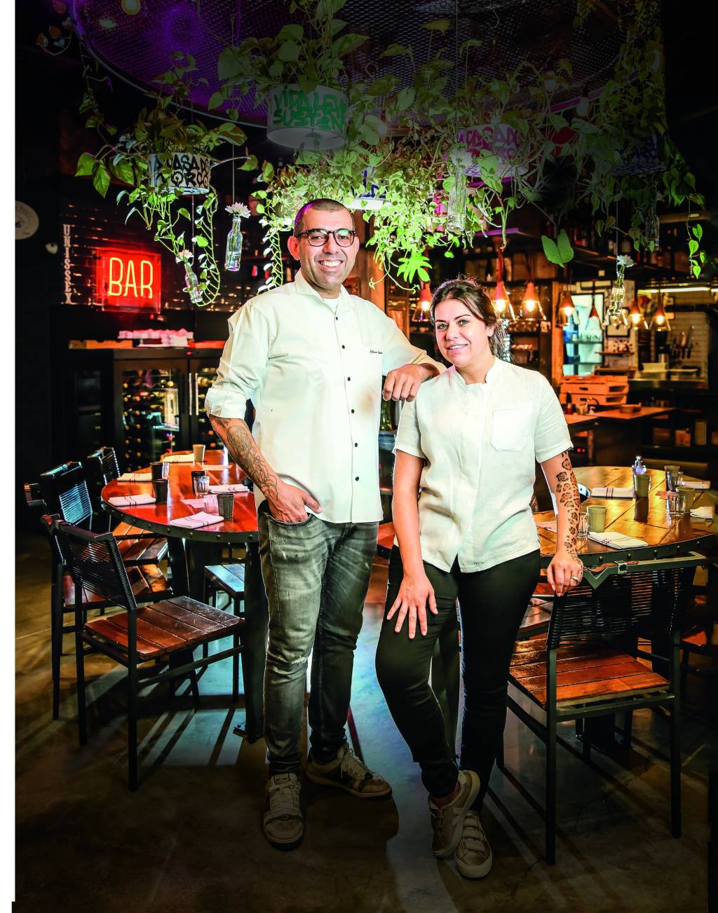 Casal branco de chefs trajados de dólmã no meio do salão de restaurante.