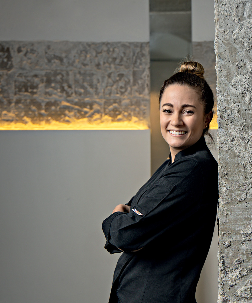 Giovanna Grossi Mulher de dólmã preto encostada em parede cinza olhando para foto sorrindo.