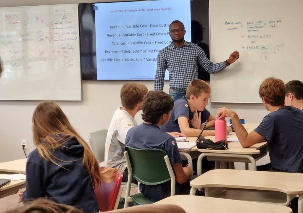 Imagem mostra sala de aula com mesas quadradas, alunos sentados em cadeiras prestando atenção em professor, que aparece ao fundo na frente de duas lousas e imagem de um projetor, com texto sobre aula de negócios em inglês