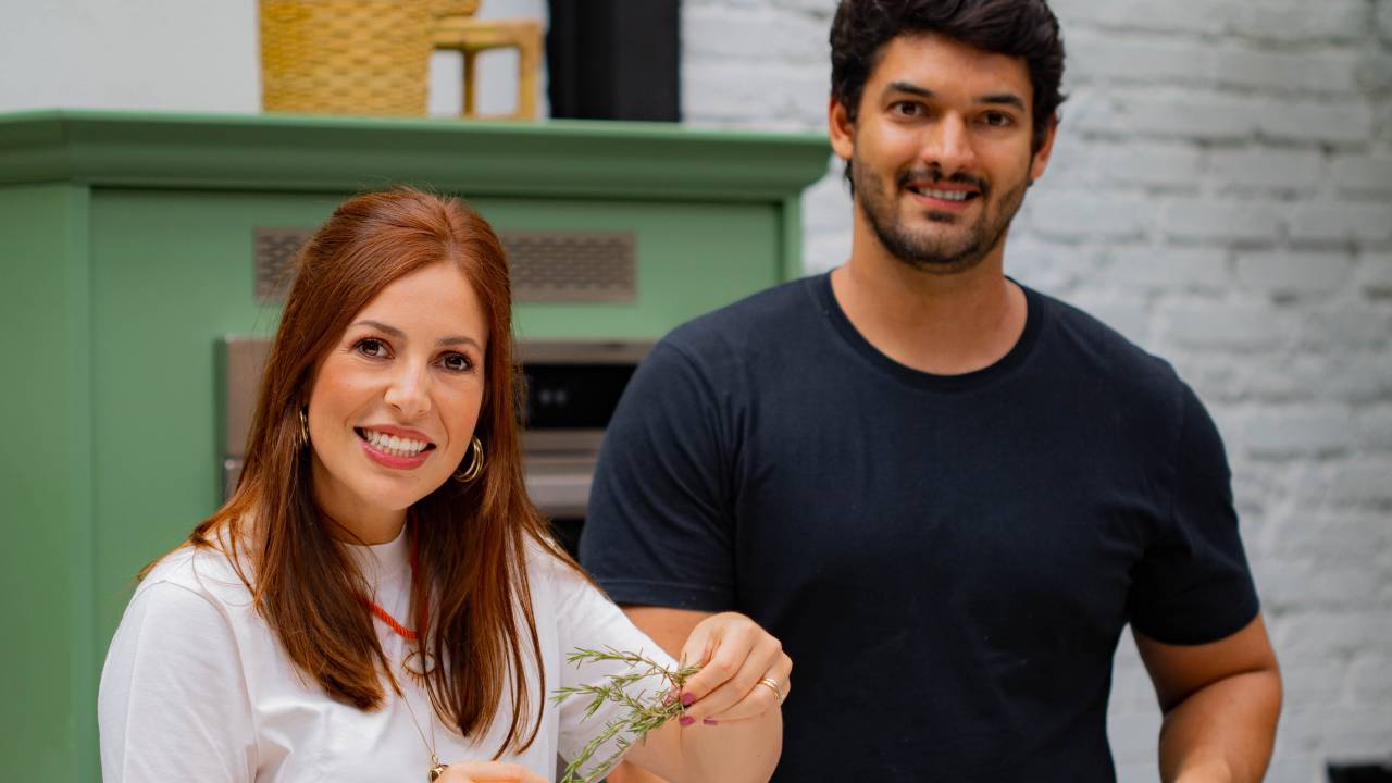 Mulher branca e ruiva posa ao lado de homem com cabelos e barba pretos. Ela veste camisa branca e ele uma camiseta preta. Ambos estão em uma cozinha, manipulando utensílios de culinária.