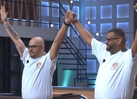 Imagem mostra Diego à esquerda e Wilson à direita, ambos de braços levantados e mãos dadas. Eles estão no cenário do Masterchef e vestem roupas de chefs de cozinha