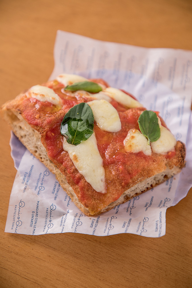 Pizza Al Taglio Margherita – Qui o Qua Fatia de pizza retangular com molho vermelho, pedaços de queijo e manjericão. Sob um prato branco em cima de uma mesa de madeira.