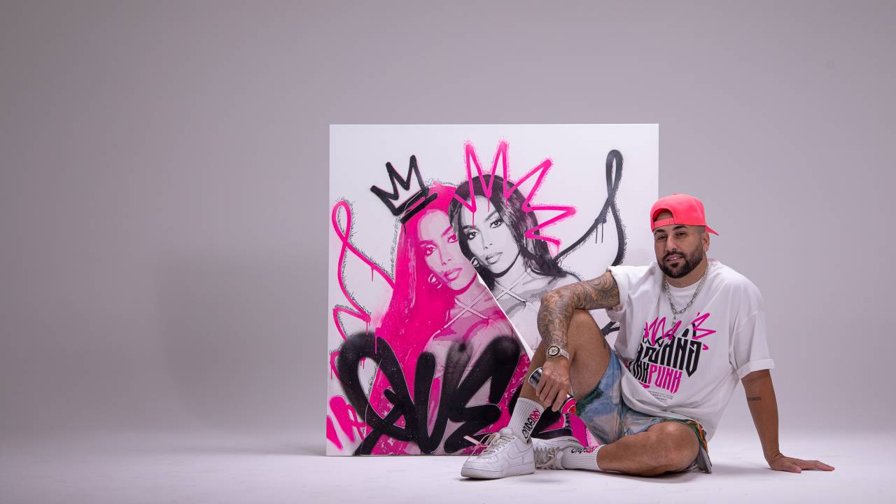 Homem de camiseta branca estampada e boné rosa virado para trás posa sentado com um dos joelhos dobrados e a mão apoiada nele. Ao seu lado, um quadro branco com grafites em rosa e preto adornam a figura da cantora Anitta. O fundo do ambiente é levemente cinza.