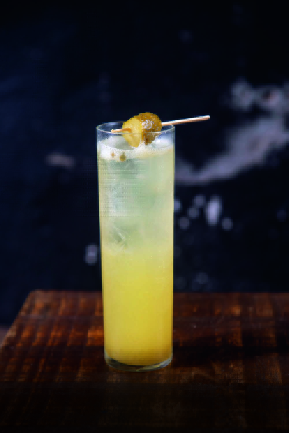 Fiíca Bar Drinque amarelado em copo longo sobre mesa de madeira.