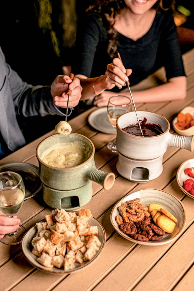 Mesa de madeira com duas panelas de fondue, uma de queijo e outra de chocolate, ao lado de cumbucas com pão cortado e frutas.