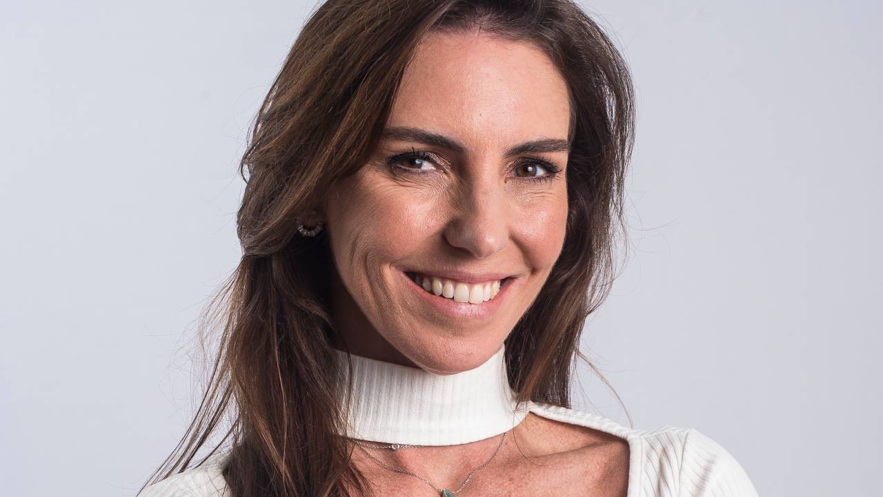 Apresentadora Glenda Kozlowski posa de braços cruzados sorrindo. Tem a pela branca, os cabelos longos lisos e escuros. Veste blusa branca com manga longa, uma fenda que exibe um pequeno pedaço do colo e tampa o pescoço.