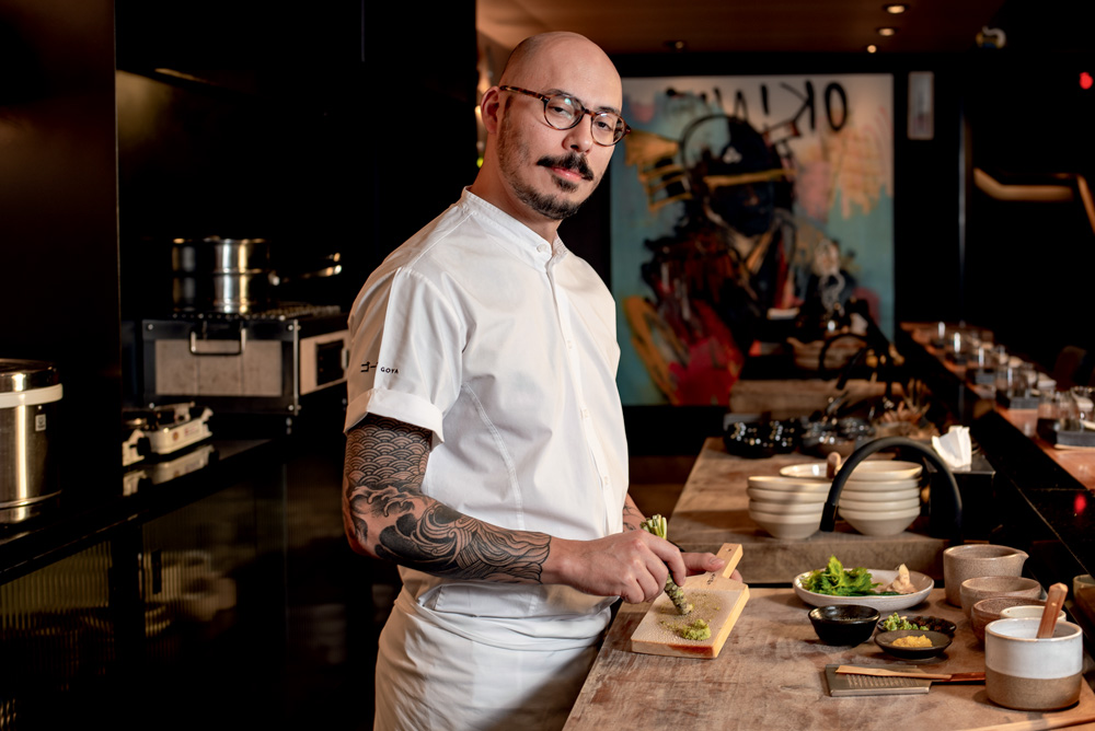 Sushiman de dólmã ralando wassabi em tábua de madeira em cozinha de restaurante.