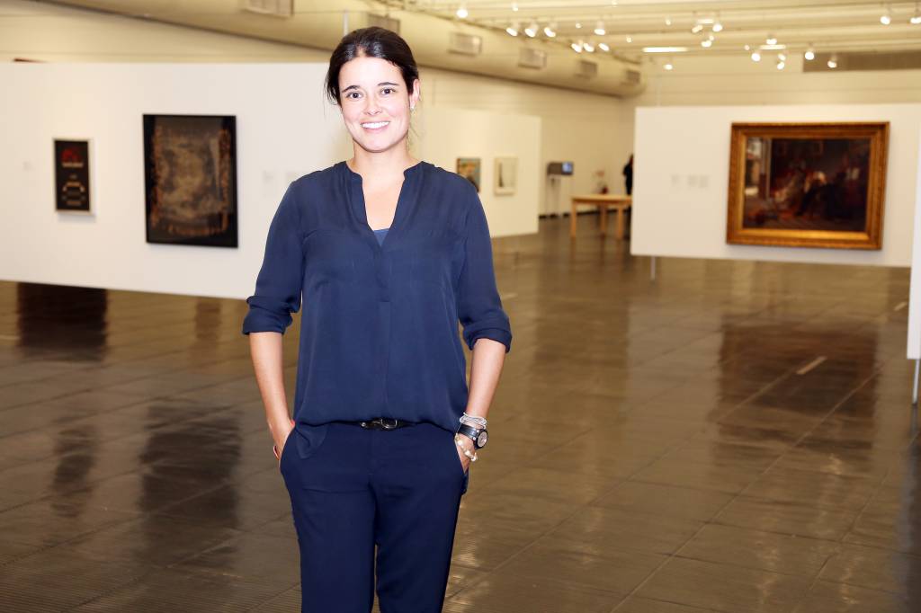 Mulher branca com cabelo preso posa com mãos no bolso em ambiente com quadros ao fundo. Veste roupa inteiramente azul.